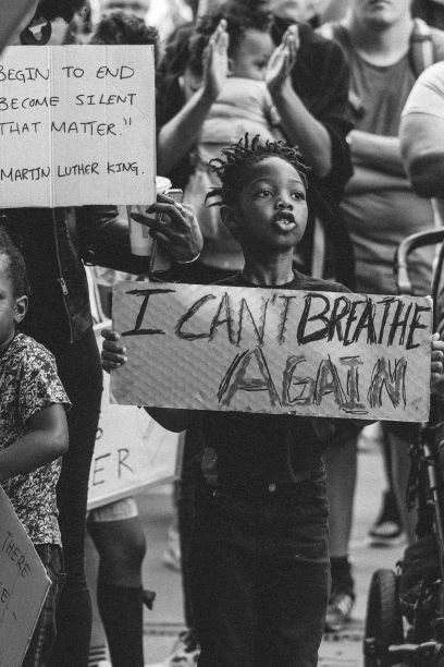 A young Black child holding a handwritten protest sign that says, "I can't breathe again."